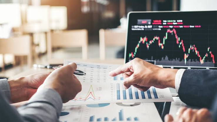 Two financial advisers reviewing investment portfolio reports and charts, with a stock market trading screen visible in the background.